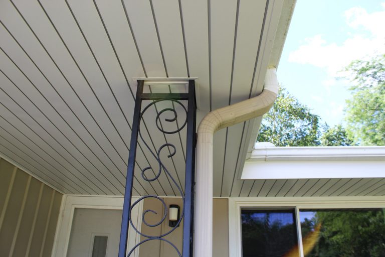Outdoor porch ceiling with a decorative metal lamp post next to a white downspout, against a backdrop of trees and sky.