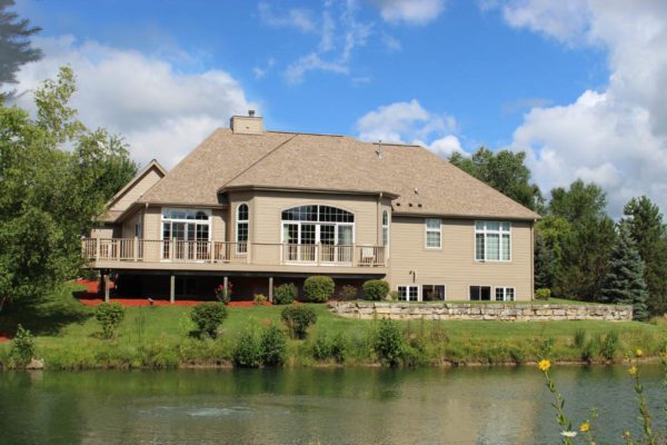 A large beige house with siding, many windows, and a deck, overlooking a pond surrounded by greenery under a blue sky with fluffy clouds.