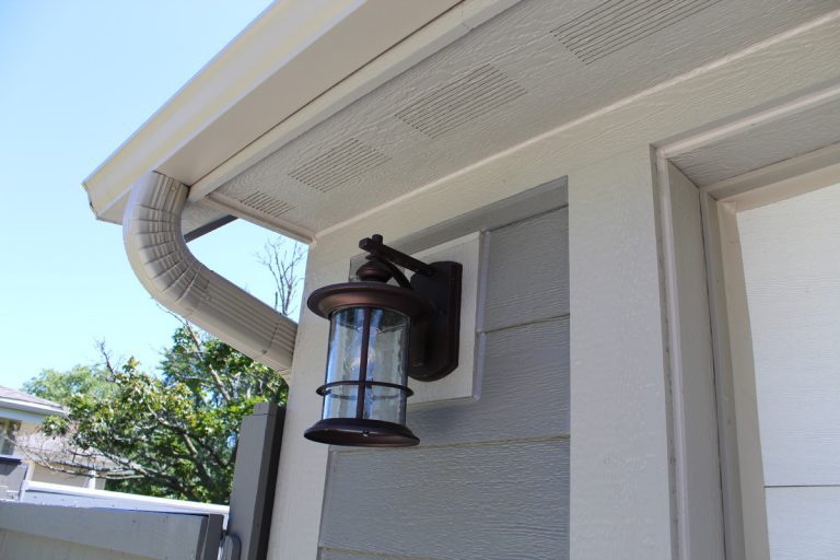 Wall-mounted outdoor lantern on a white house with gray siding under a clear blue sky.