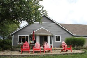 A suburban gray house with a white door, surrounded by greenery, featuring a front patio with four red Adirondack chairs.