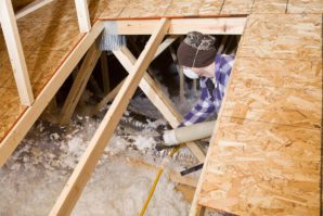A worker insulating the floor of an attic using a hose to apply spray foam, surrounded by wooden beams and plywood flooring.