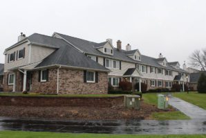 A row of multi-unit residential buildings with brick and siding exteriors on a rainy day, featuring wet roads and lawns.