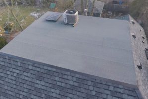 Aerial view of a flat rooftop with a central air conditioning unit, surrounded by a shingled sloping roof, overlooking a backyard.