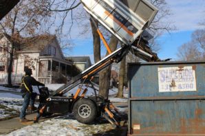 A person operates a small, wheeled mechanical lift to dump a load of construction debris into a dumpster on a snowy street.