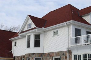 A white house with red shingle siding and a stone foundation, featuring a balcony and a dormer window.