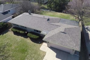 Aerial view of a one-story suburban house with a gray shingled roof, surrounded by green lawn and neighboring homes.