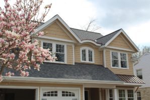 A suburban house with beige siding, a dark gray roof, and a blooming magnolia tree to the left.