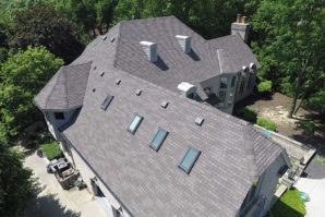 Aerial view of a large gray-roofed house surrounded by dense green trees on a sunny day.