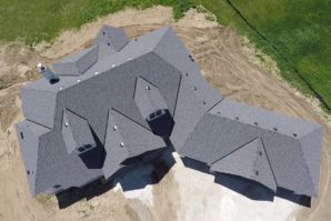 Aerial view of a large, complex roof on a newly constructed house featuring multiple angles and dormer windows, surrounded by a dirt landscape.