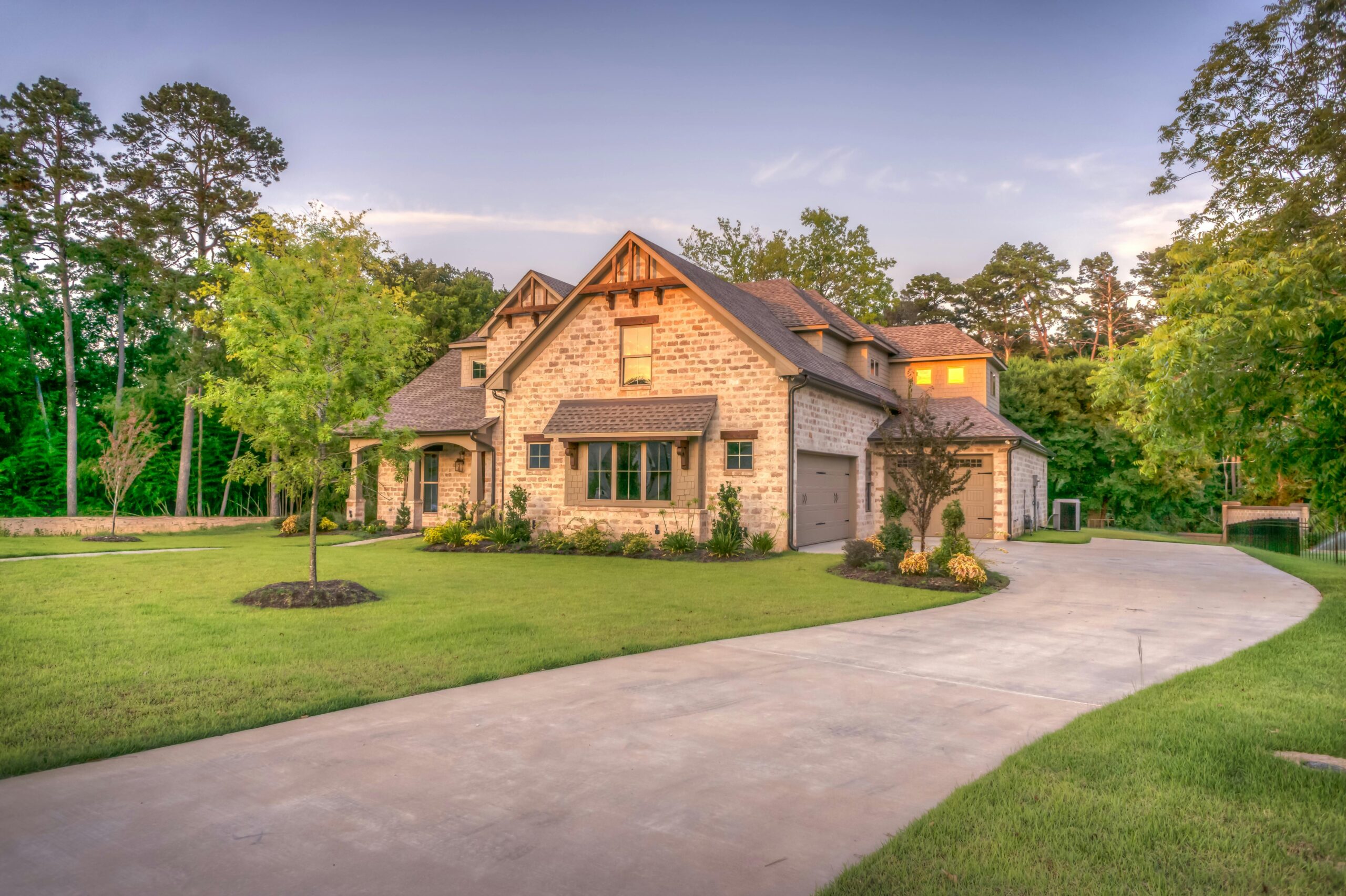 A two-story stone house with siding, a large driveway, manicured green lawn, and landscaped garden, surrounded by tall trees under a clear sky at sunset.